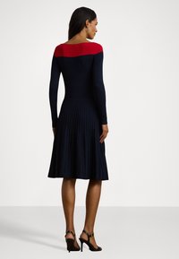 Vestido de malha com mangas longas; corpo azul-marinho e parte superior vermelha. Design de saia plissada. Usado com sandálias de salto alto pretas.