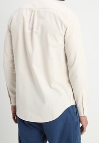 Beige overhemd met lange mouwen, een puntsak, een rugpand en een gebogen zoom. Lichtjes textielachtig stof met knoopmanchetten en een open placket.