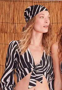 Femme aux cheveux ondulés portant un foulard à rayures zèbre noir et blanc et un crop top noué assorti, debout devant un fond en bambou.