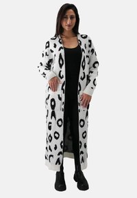 Langer, weißer Cardigan mit schwarzem Leopardenmuster, mit gerippten Bündchen und Taschen, über einem schwarzen Outfit getragen, detaillierte Textur sichtbar.