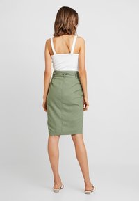 Jupe en lin vert olive, longueur genoux, taille haute, fente arrière et passants de ceinture, associée à un débardeur blanc. Design simple, sans motifs.