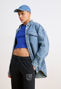 Džinsi jaka ar pogām, zila pieguļošā virsotne, melni sporta bikses ar Nike logo, un gaiši zila cepure ar logo, akcentējot ikdienišķo ielu stilu.
