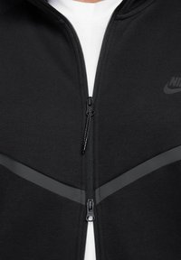 Melnas ar rāvējslēdzēju aizveramo jaku, kurai ir atstarojoši akcenti, gluda tekstūra un izteikts rāvējslēdzējs. Nike logo redzams augšējā kreisajā pusē.