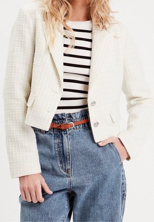 Femme portant un blazer court blanc texturé, un top rayé noir et blanc, un jean bleu taille haute et une ceinture marron, main dans la poche.