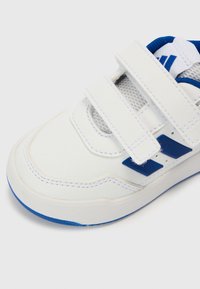 Valkea urheilukenkä sinisin korostuksin, sileää synteettistä materiaalia, tarrakiinnitykset, rei'itetty verkkokieleke ja kuvioitu sininen pohja.