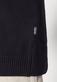 Maglione nero lavorato a maglia con superficie texturizzata, orlo a coste e etichetta laterale. Il materiale sembra spesso e resistente, adatto per la stagione più fresca.