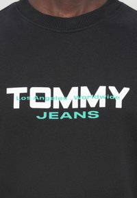 Μαύρο φούτερ με ριμπ λαιμόκοψη, με έντονη λευκή και τιρκουάζ γραφή: «TOMMY» και «JEANS», καθώς και «Los Angeles, Worldwide» με μικρότερη γραμματοσειρά.