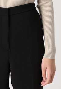 Pantalones negros ajustados con una textura suave y una cinturilla estructurada, combinados con una manga de suéter ligero acanalado visible en la parte superior.