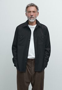 Camicia nera con bottoni e due tasche sul petto, indossata sopra una t-shirt bianca. Pantaloni marroni con pieghe. Tessuto movimentato, vestibilità rilassata.