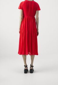 Abito rosso plissettato con maniche corte, un corpetto aderente e una gonna fluente. Sandali neri con tacco spesso e cinturini alla caviglia completano il look.