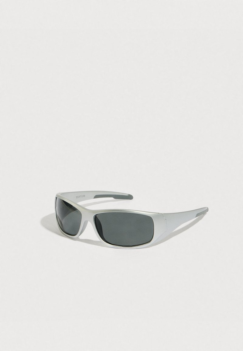 Γυαλιά ηλίου με ασημένιο πλαστικό σκελετό, σκουρόχρωνους γκρι φακούς και κυρτούς βραχίονες. Οι φακοί έχουν ελαφρώς περιμετρικό σχεδιασμό για κάλυψη.