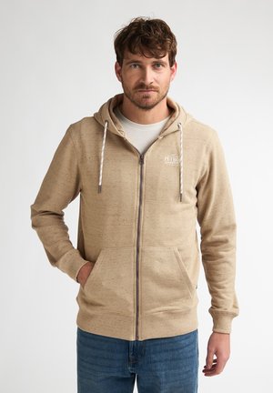 Sweat à capuche beige à fermeture éclair, en tissu doux, avec une fermeture éclair frontale, une capuche à cordon de serrage et des poches kangourou. Logo texte sur la poitrine.