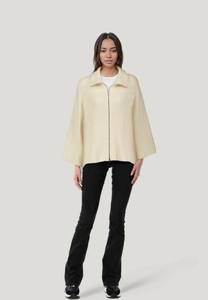Cremfarbener gerippter Zip-Cape mit Kragen, mit weiten Ärmeln, kombiniert mit schwarzen Schlagjeans und schwarzen Turnschuhen.