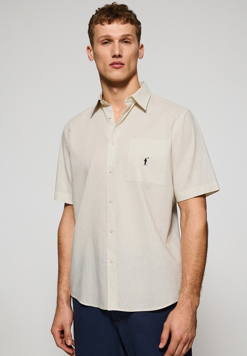 Korte mouwen, licht beige shirt gemaakt van stof met een subtiele textuur. Bevat een borstzak en een klein zwart logo dat erop geborduurd is.