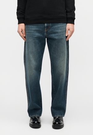 Osoba ubrana w ciemne dżinsy o prostych nogawkach, czarne błyszczące skórzane buty oraz czarną bluzkę z długim rękawem, stojąca z rozluźnionymi rękami.