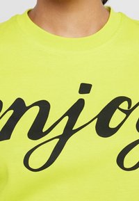 Sudadera amarilla neón con un gran texto en cursiva negra que dice "enjoy" en la parte delantera, con un cuello redondo y una textura de tela suave.