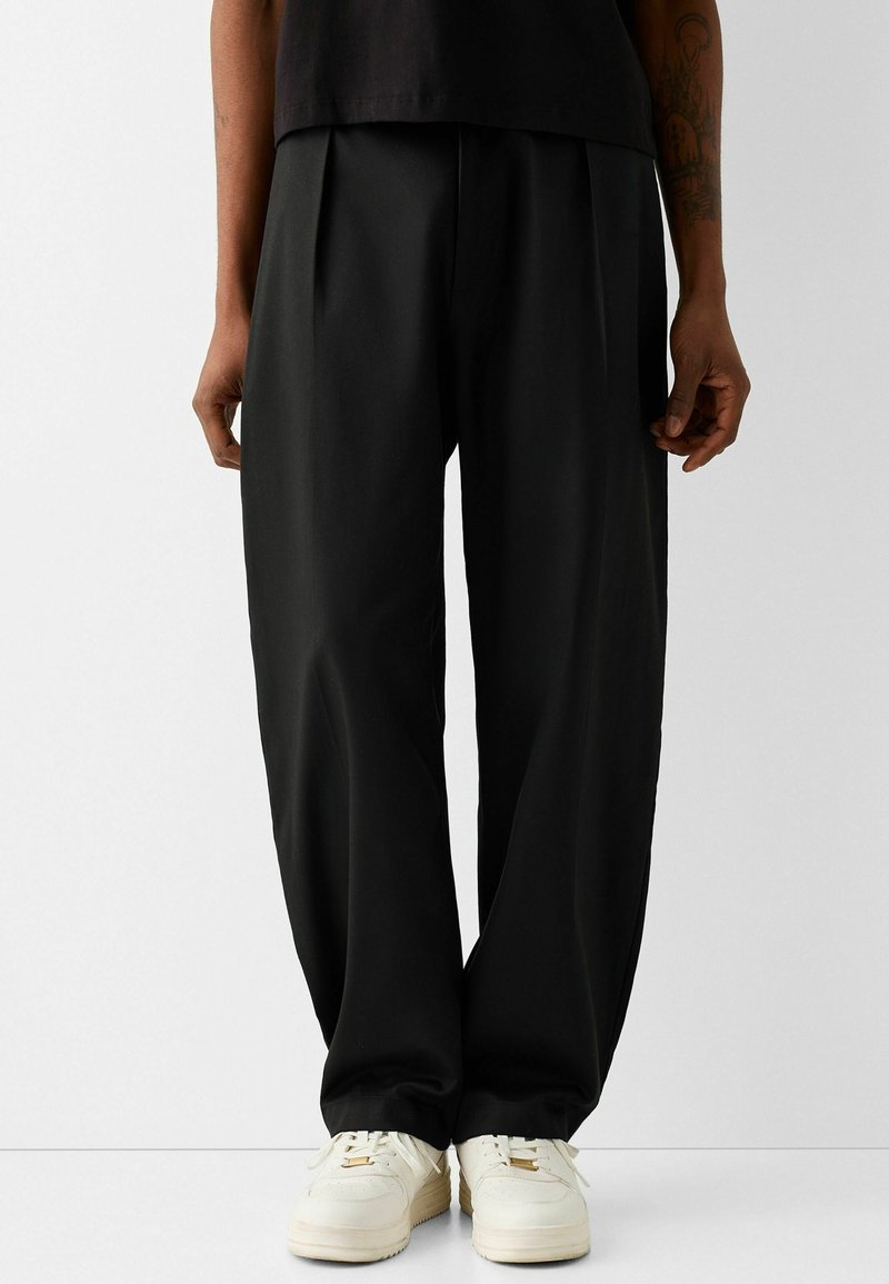 Personne portant un pantalon noir ample, un haut noir et des baskets blanches, debout devant un fond clair uni.