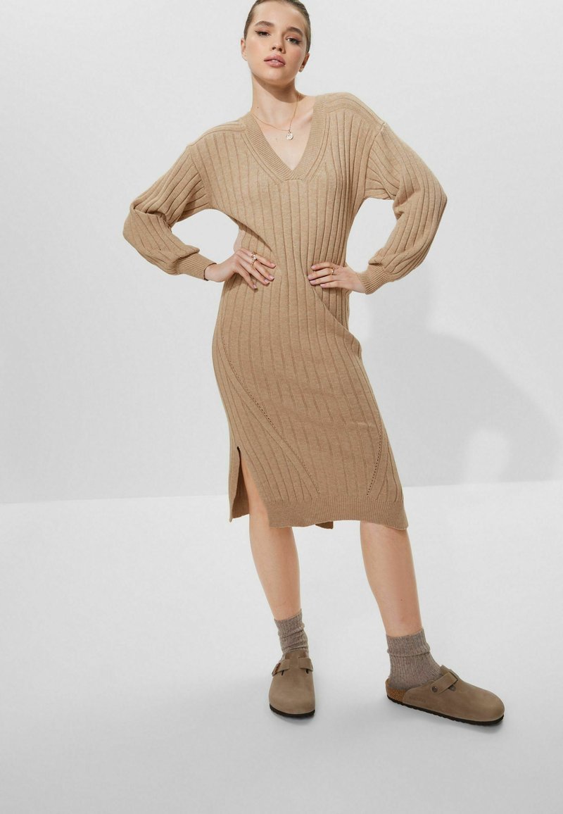 Robe en tricot beige avec texture côtelée, décolleté en V et manches longues ; dotée d'une fente latérale. Portée avec des sabots marron et des chaussettes gris à la cheville.