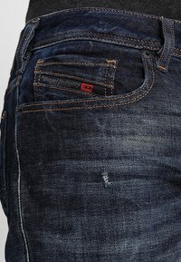 Tmavě modré džíny z denimu s oranžovým prošíváním, malou červenou nášivkou na zadní kapse a s distressed detailem blízko lemu.