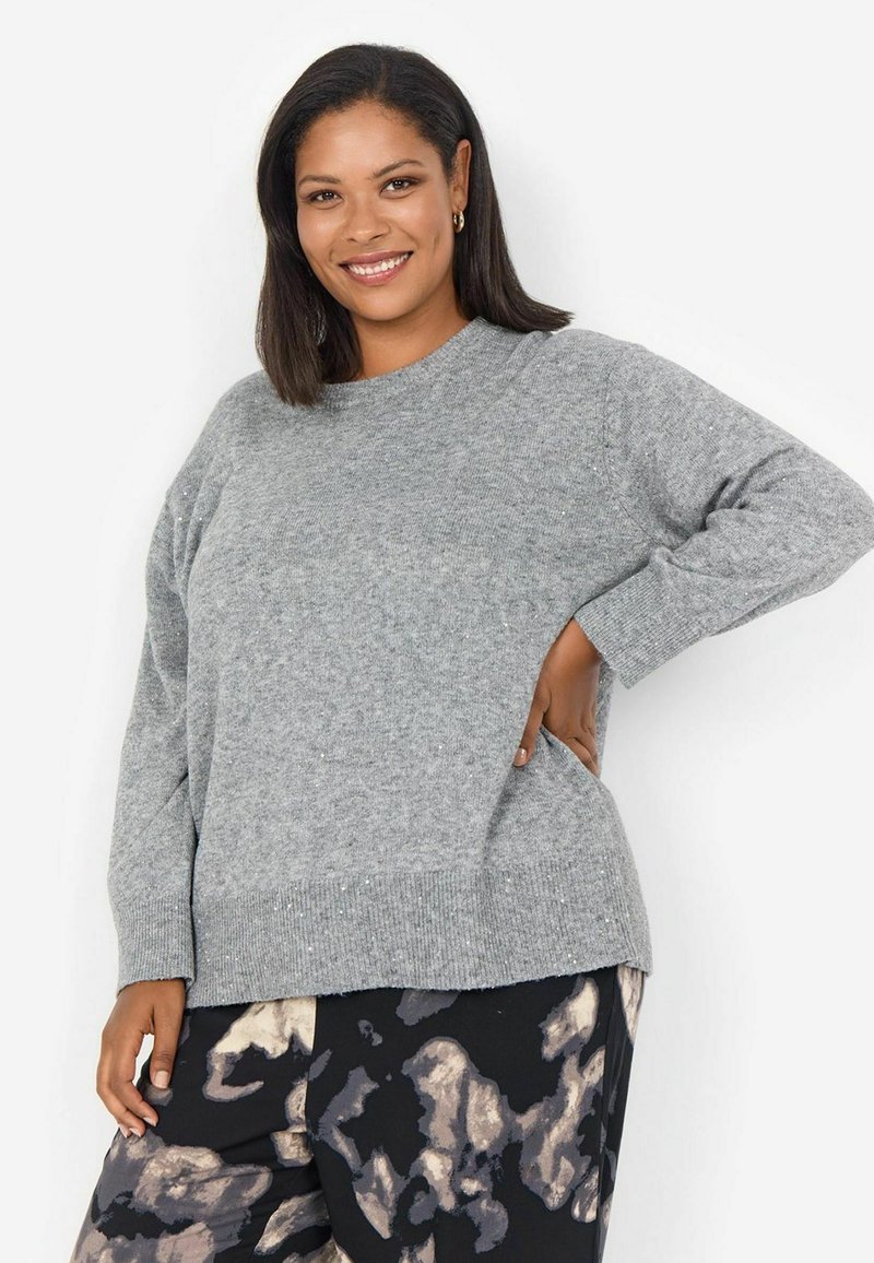 Pull en tricot gris clair à col rond, avec un ourlet côtelé et des manches longues ; associé à un pantalon sombre à motifs avec des formes abstraites.