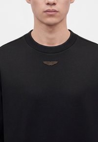 Gros plan d'une personne portant un t-shirt noir uni à col rond avec un petit emblème foncé en forme d'aile au centre de l'encolure avant.