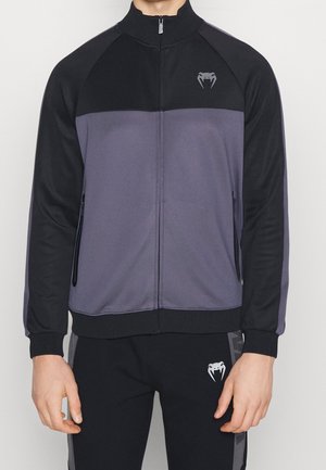 Man wearing black and gray full-zip jacket and black pants with snake fang logo on chest and upper thigh.