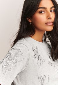 Vrouw met lang donker haar, gekleed in een lichtgrijze shirt met zilveren pailletjes bloemenborduurwerk en kleine zilveren creolen.