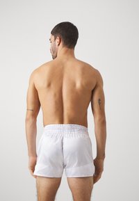 White satin swim shorts with an elastic waistband, smooth texture, and a mid-thigh cut, worn by a male model viewed from the back.
