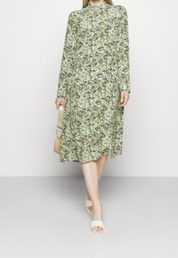 Grünblumiges, langärmliges Kleid mit hohem Ausschnitt und einer fließenden Silhouette. Der Stoff zeigt zahlreiche kleine rosa und weiße Blumen.
