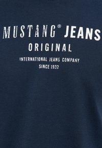 Navy blue cotton t-shirt featuring bold white text: "MUSTANG JEANS ORIGINAL" and "INTERNATIONAL JEANS COMPANY SINCE 1932." Smooth texture.