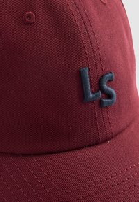 Burgundi sapka textúrált pamut anyagból. Elöl hímzett fekete "LS" logóval, varrott részletekkel és szellőző szemekkel rendelkezik.