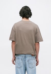 Person with short wavy hair wearing a loose brown t-shirt and light blue jeans, standing facing away against a plain white background.