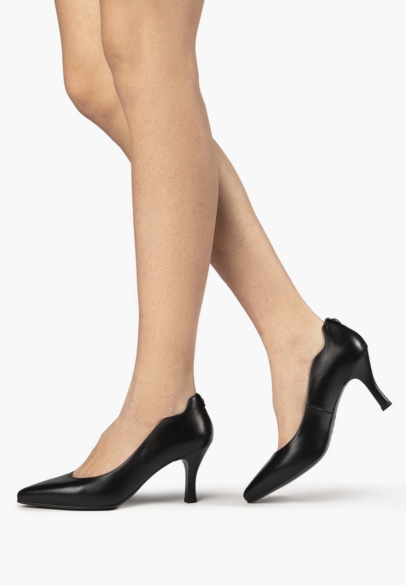 Jambes portant des escarpins noirs en cuir à bout pointu avec des talons fins de hauteur moyenne, présentés sur un fond blanc uni.