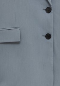 Section de blazer en tissu bleu présentant une texture lisse, deux boutons noirs et une petite poche à rabat sur le côté gauche.