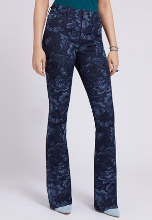 Kobieta ubrana w ciemnoniebieskie dzwony w kwiatowy wzór, w połączeniu z jasnoniebieskimi, szpiczastymi butami, stojąca na tle jednolitego tła.