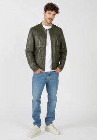 Grüne Lederjacke mit zwei Brusttaschen, runder Halslinie und Reißverschluss, kombiniert mit hellblauen Jeans und weißen Sneakers.