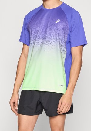 Mann, der ein kurzärmeliges Sportshirt mit lila-grünem Farbverlauf und schwarzen Shorts trägt, steht vor einem einfarbigen hellen Hintergrund.