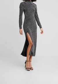 Žena v dlouhých rukávech, oblečená v černobílé skvrnité šaty s vysokým rozparkem, v kombinaci s černými sandály na vysokém podpatku s kotníkovým páskem, stojící na jednobarevném pozadí.