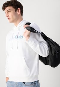 Biała bluza z kapturem i kieszenią z przodu, z jasnoniebieskim logo. Czarny plecak z zamkiem, noszony na jednym ramieniu. Dżinsy o luźnym kroju.