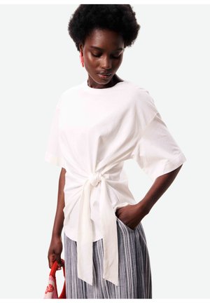 Femme aux cheveux courts et bouclés portant un haut blanc surdimensionné noué à l'avant, un pantalon large rayé, tenant un sac rouge et blanc, regardant vers le bas.