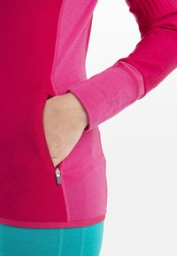 Main dans la poche zippée d'une veste de sport rose à blocs de couleurs, portée avec un pantalon turquoise, présentée sur un fond blanc uni.
