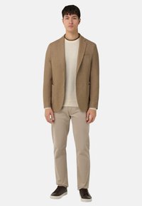 Beżowa marynarka z wyciętymi klapami, w zestawieniu z jasnym swetrem i beżowymi spodniami. Dopełnieniem stroju są brązowe trampki. Gładka faktura tkaniny.
