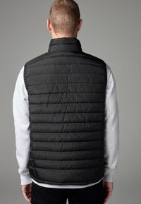 Chaleco de abrigo negro con textura acolchada, cuello alto y cierre de cremallera, llevado sobre un sudadera gris. La parte trasera presenta detalles de costura horizontal.