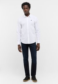 Mężczyzna stojący w białej koszuli zapinanej na guziki z logo, ciemnoniebieskich dżinsach i czarnych trampkach z gumową podeszwą na jednolitym tle.