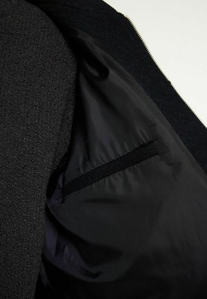 Schwarze Jacke mit glattem Innenfutter und strukturiertem Obermaterial, ausgestattet mit einer versteckten Tasche, die in die Seitennähten eingenäht ist.