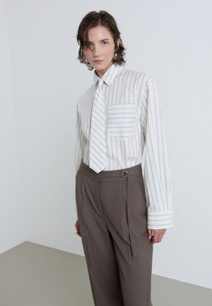 Personne portant une chemise blanche à rayures verticales avec une cravate assortie et un pantalon marron taille haute avec une ceinture latérale, debout devant un fond uni.