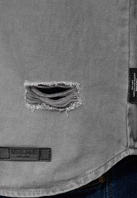 Grijs denim overhemd met een versleten scheur met rafelige randen en een leren label met "INDICODE EST. 1993" aan de zoom.