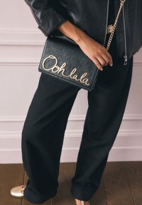 Juodos odos delninė su auksine kursyvine užrašu „Ooh la la“; stačiakampio formos su grandinėlės dirželiu; rankoje laiko asmuo, dėvintis juodas kelnes.