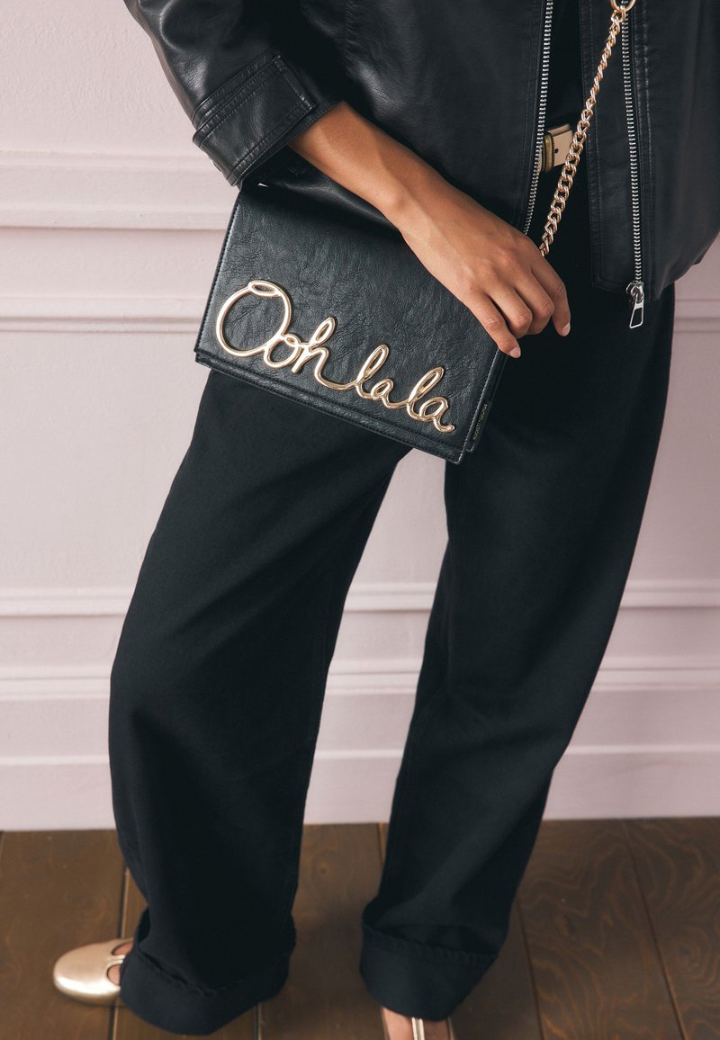 Juodos odos delninė su auksine kursyvine užrašu „Ooh la la“; stačiakampio formos su grandinėlės dirželiu; rankoje laiko asmuo, dėvintis juodas kelnes.