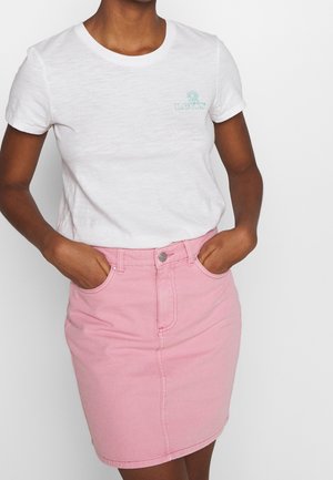 Personne portant un t-shirt blanc à manches courtes avec un petit logo floral vert et une jupe en denim rose taille haute, les mains dans les poches.